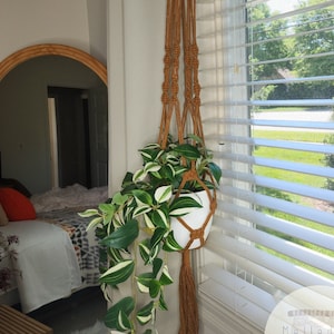 May include: A macrame plant hanger with a white pot and a trailing plant with green and white leaves. The hanger is made of natural brown cord and has a tassel at the bottom. The plant hanger is hanging in a window with white blinds. The text "Melled It Handmade macrame arts and decor..." is visible in the lower right corner of the image.