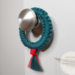 May include: A circular macrame ring in deep teal, featuring a knotted texture and a small decorative tassel at the bottom with red and teal threads. The ring is suspended from a brushed silver metal door knob on a white door. A logo reads Melled it.