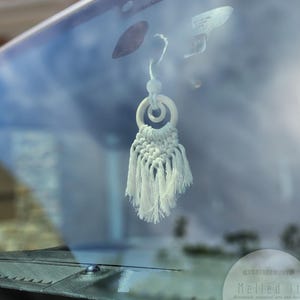 May include: White macrame car charm with a wooden ring and a white tassel hanging from the rearview mirror of a car. The charm is hanging in front of a blue sky and a house.