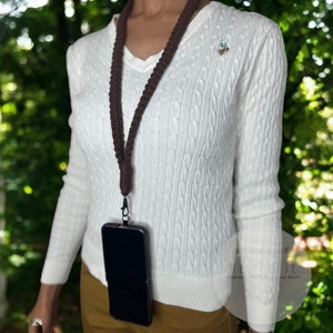 May include: A brown crocheted phone strap with a black phone case attached. The phone case has a black screen and a black border. The strap is worn by a person wearing a white sweater.