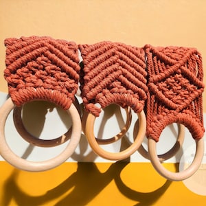 May include: Three terracotta-colored macrame towel rings with wooden rings. The macrame is made with a diamond pattern.
