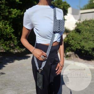 May include: A gray macrame crossbody phone bag with a matching strap. The bag has a small square pocket and a woven design. The phone is visible in a clear case. The strap is worn across the body.