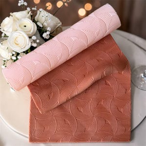 May include: A rolled pink silicone baking mat and a flat terracotta-colored mat with a repeating curved pattern. The mats are designed for baking and are shown with a bouquet of white roses in the background.