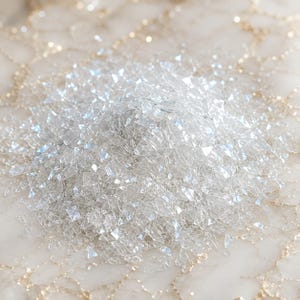 May include: A pile of small, clear, iridescent glass shards with sharp, angular edges. The fragments are translucent and reflect light, creating a shimmering effect across the collection of tiny, jagged pieces of crushed glass material.