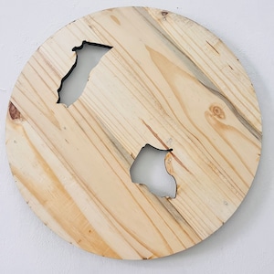 Puede incluir: Una placa redonda de madera con dos recortes en forma de continentes. La placa es de color marrón claro con veta de madera natural.