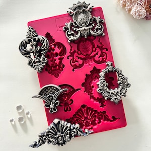 May include: A red silicone mold with six different ornate designs. The designs are in the shape of a crown, a swirl, a circle, a fan, a flower, and a scroll. The mold is used for making decorative cake toppers or other edible decorations.