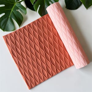 Peut inclure: Un tapis texturé de couleur corail et un tapis rose enroulé avec un motif de vagues répétées. Les tapis sont probablement destinés à l'artisanat ou à la pâtisserie, avec un design décoratif. L'image est prise d'en haut, avec une feuille verte en arrière-plan.