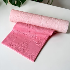 Puede incluir: Dos rodillos de silicona rosa con un patrón de hojas. Uno está enrollado y el otro plano. Los rodillos están diseñados para hornear y crear superficies texturizadas en masa o fondant.