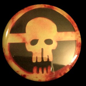 Puede incluir: Un botón redondo con un diseño de calavera y huesos cruzados en negro y naranja. La calavera es amarilla y los huesos cruzados son naranjas. El fondo es de color naranja oscuro.