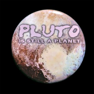 Puede incluir: Un botón redondo con una imagen de Pluton. El texto "Plutón sigue siendo un planeta" está escrito en letras rosas en el botón.