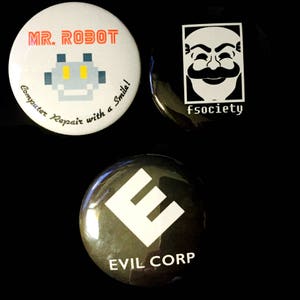 Puede incluir: Tres botones redondos con diferentes diseños. El primer botón es blanco con texto rojo y negro que dice "MR. ROBOT" y una imagen pixelada de un robot con ojos amarillos. El texto debajo del robot dice "Computer Repair with a Smile!" El segundo botón es negro con texto blanco que dice "fsociety" y una imagen blanca de un hombre con barba y una expresión seria. El tercer botón es verde oscuro con una letra "E" blanca y texto blanco que dice "EVIL CORP".