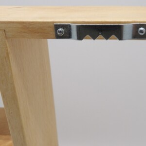 Puede incluir: Primer plano de una estanter&iacute;a de madera con un soporte de suspensi&oacute;n met&aacute;lico fijado en la parte superior. El soporte tiene un borde dentado y est&aacute; asegurado con dos tornillos.
