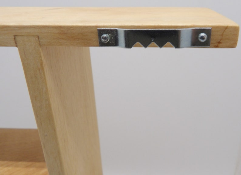 Puede incluir: Primer plano de una estanter&iacute;a de madera con un soporte de suspensi&oacute;n met&aacute;lico fijado en la parte superior. El soporte tiene un borde dentado y est&aacute; asegurado con dos tornillos.
