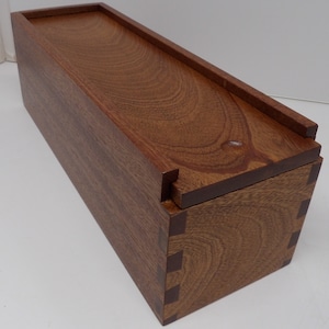 May include: A wooden box with a hinged lid. The box is made of dark brown wood with a natural grain pattern. The lid is slightly raised and has a small lip around the edge. The box is closed.