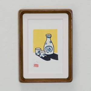 May include: A framed print featuring a block-print style illustration of a white sake bottle and two small cups with blue floral patterns against a yellow background. The bottle displays the blue Japanese kanji character for sake. A red stamp is visible.