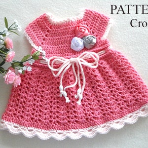 May include: A pink crochet baby dress with a white crocheted trim and a white ribbon tied at the waist. The dress has a flower embellishment on the bodice.