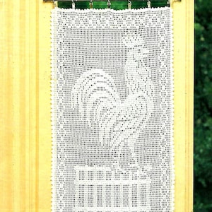 May include: White crocheted curtain with a rooster design hanging on a wooden door frame.