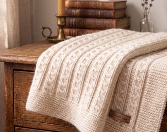 May include: A cream-colored knitted blanket featuring a textured cable knit pattern and a garter stitch border. A brown rectangular fabric label is attached to the corner, with the text Knit with Love printed in white lettering.