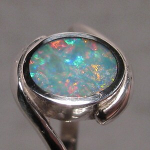 May include: A silver ring with an oval opal gemstone. The opal exhibits a colourful play of light, with hues of blue, green, red, and yellow. The ring's band has a distinctive, swirling design, enhancing its elegant appearance.