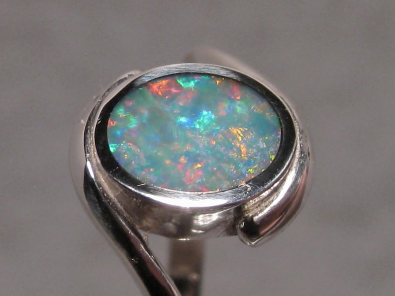 May include: A silver ring with an oval opal gemstone. The opal exhibits a colourful play of light, with hues of blue, green, red, and yellow. The ring's band has a distinctive, swirling design, enhancing its elegant appearance.