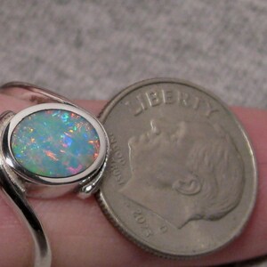 May include: A silver ring featuring an oval opal gemstone. The opal exhibits a vibrant play of colour, including blues, greens, and oranges. The ring is next to a US dime coin for size comparison.