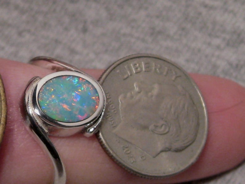 May include: A silver ring featuring an oval opal gemstone. The opal exhibits a vibrant play of colour, including blues, greens, and oranges. The ring is next to a US dime coin for size comparison.