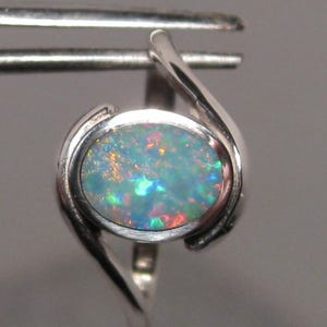 May include: A silver ring with an oval opal gemstone. The opal exhibits a colourful play-of-colour with shades of blue, green, and red. The ring has a contemporary, minimalist design.