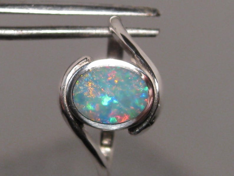 May include: A silver ring with an oval opal gemstone. The opal exhibits a colourful play-of-colour with shades of blue, green, and red. The ring has a contemporary, minimalist design.