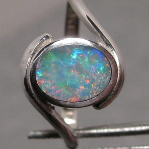 May include: A silver ring with an oval opal gemstone. The opal exhibits a vibrant play of colour with blues, greens, and reds. The ring's design has a modern, flowing shape, with the band curving around the gemstone.