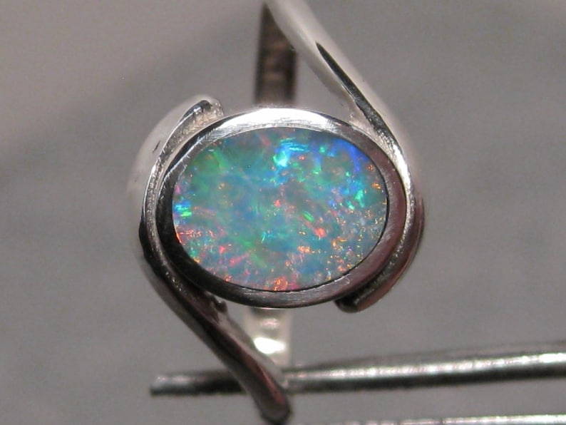 May include: A silver ring with an oval opal gemstone. The opal exhibits a vibrant play of colour with blues, greens, and reds. The ring's design has a modern, flowing shape, with the band curving around the gemstone.