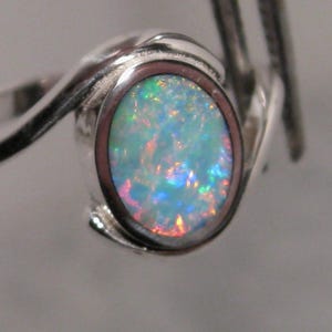 May include: A silver ring with an oval opal gemstone. The opal exhibits a vibrant play of colour with blues, greens, and reds. The ring's band has a swirling design, enhancing its elegant appearance.