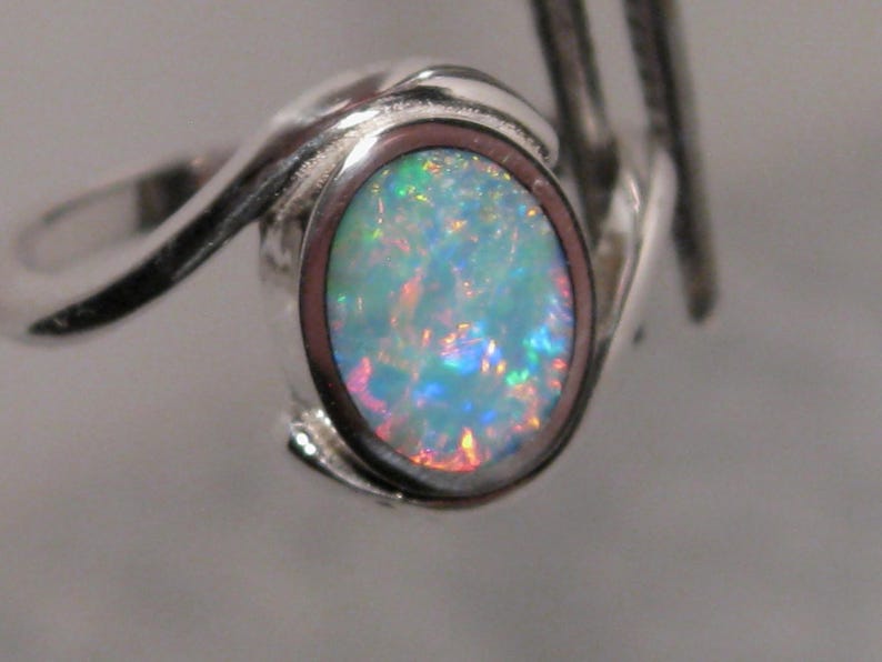 May include: A silver ring with an oval opal gemstone. The opal exhibits a vibrant play of colour with blues, greens, and reds. The ring's band has a swirling design, enhancing its elegant appearance.