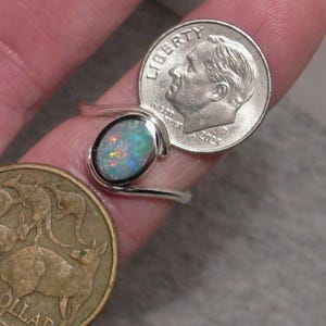 May include: A silver ring featuring an oval opal gemstone. The opal shows a mix of blue, green, and orange hues. The ring is next to a U.S. dime and an Australian one-dollar coin.