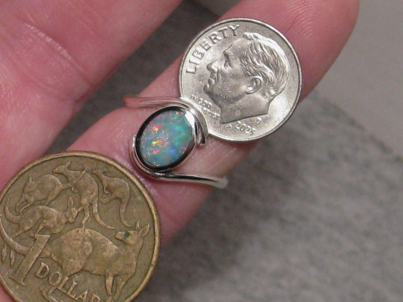 May include: A silver ring featuring an oval opal gemstone. The opal shows a mix of blue, green, and orange hues. The ring is next to a U.S. dime and an Australian one-dollar coin.