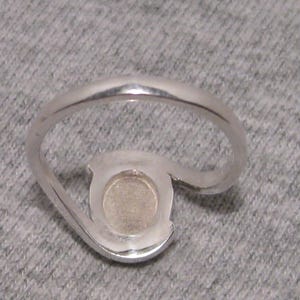 May include: A silver ring with a distinctive, open design. It has a circular setting with a light-coloured centre stone. The band curves around the setting, creating an elegant, modern appearance. The ring is made of silver.