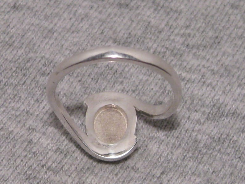 May include: A silver ring with a distinctive, open design. It has a circular setting with a light-coloured centre stone. The band curves around the setting, creating an elegant, modern appearance. The ring is made of silver.