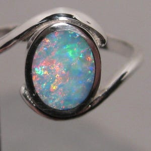 May include: A silver ring featuring an oval opal gemstone. The opal exhibits a vibrant play of colour, with flashes of blue, green, and red. The ring's band has a flowing, curved design, enhancing its elegant appearance.