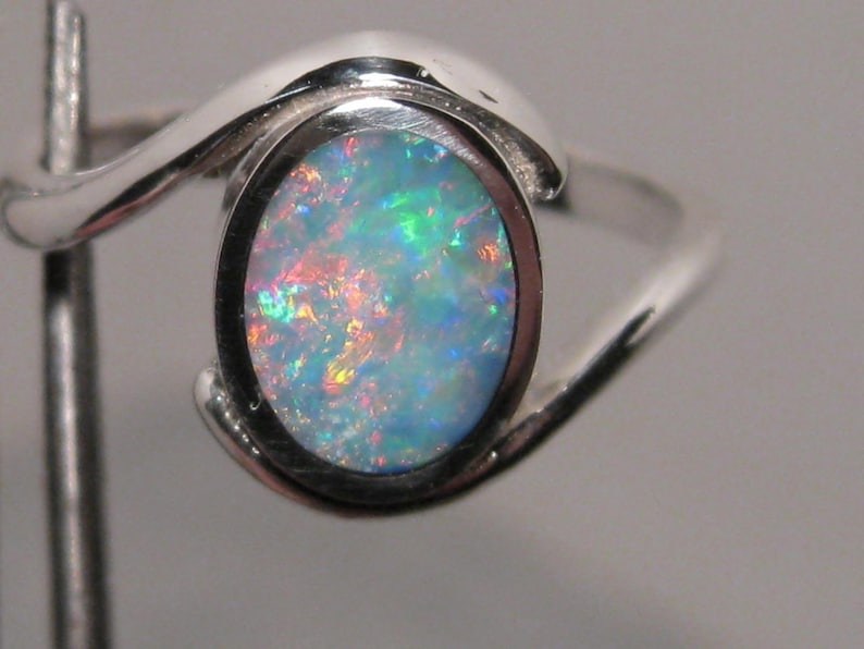 May include: A silver ring featuring an oval opal gemstone. The opal exhibits a vibrant play of colour, with flashes of blue, green, and red. The ring's band has a flowing, curved design, enhancing its elegant appearance.