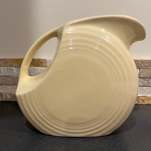 May include: A cream-colored ceramic pitcher with a rounded shape and a wide, flared spout. The pitcher has a smooth, glossy finish and features a series of concentric rings around the body.