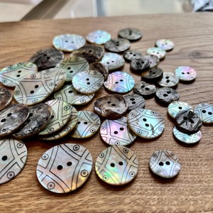 May include: A selection of mother-of-pearl buttons in different sizes and patterns. The buttons have iridescent and dark brown hues, with some showing a geometric design. Each button has two holes for sewing.