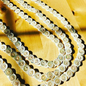 May include: Multiple strands of white button garland are displayed diagonally on a light wooden surface. The round buttons, each with four holes, have a pearlescent finish. The garland is composed of small, closely-set buttons.
