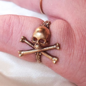 May include: A gold skull and crossbones ring. The ring is worn on a finger.