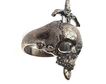 May include: A silver-toned ring featuring a skull design with a sword piercing the top. The skull has detailed features, including eye sockets and a lower jaw. The ring is set against a white background.