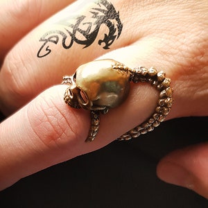 May include: A gold skull ring with a detailed design of tentacles wrapping around the skull. The ring is worn on a finger with a black dragon tattoo.