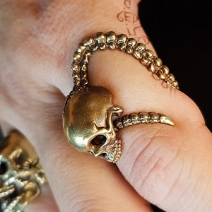 May include: A gold skull ring with a detailed design. The ring is worn on a finger and features a skull with a long, curved horn.