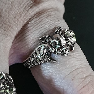 Op de afbeelding: Een zilveren ring met een schedel en vleugels ontwerp. De schedel heeft rode ogen.