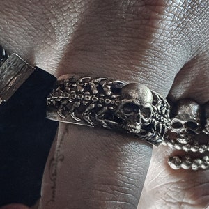 May include: A silver-toned skull ring and bracelet set. The ring features multiple small skulls, while the bracelet has a skull and a rectangular clasp. The jewelry is worn on a hand.