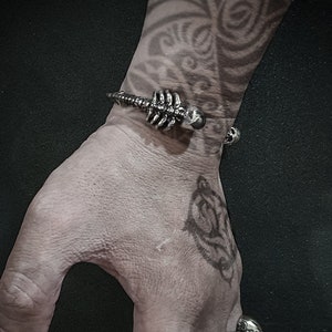 Könnte beinhalten: Zwei silberne Armbänder in Schädelform. Das Armband am Handgelenk ist ein Skelettdesign mit einem Schädel am Ende. Das Armband am Finger ist ein Ring mit mehreren Schädeln.