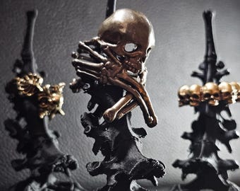 May include: Three decorative sculptures featuring a skull and rib cage design. The skulls are bronze-coloured, and the structures are black. The sculptures are displayed on a dark grey surface, creating a gothic aesthetic.
