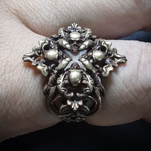 May include: A gold-toned ring with a detailed design featuring multiple skulls and intricate scrollwork.